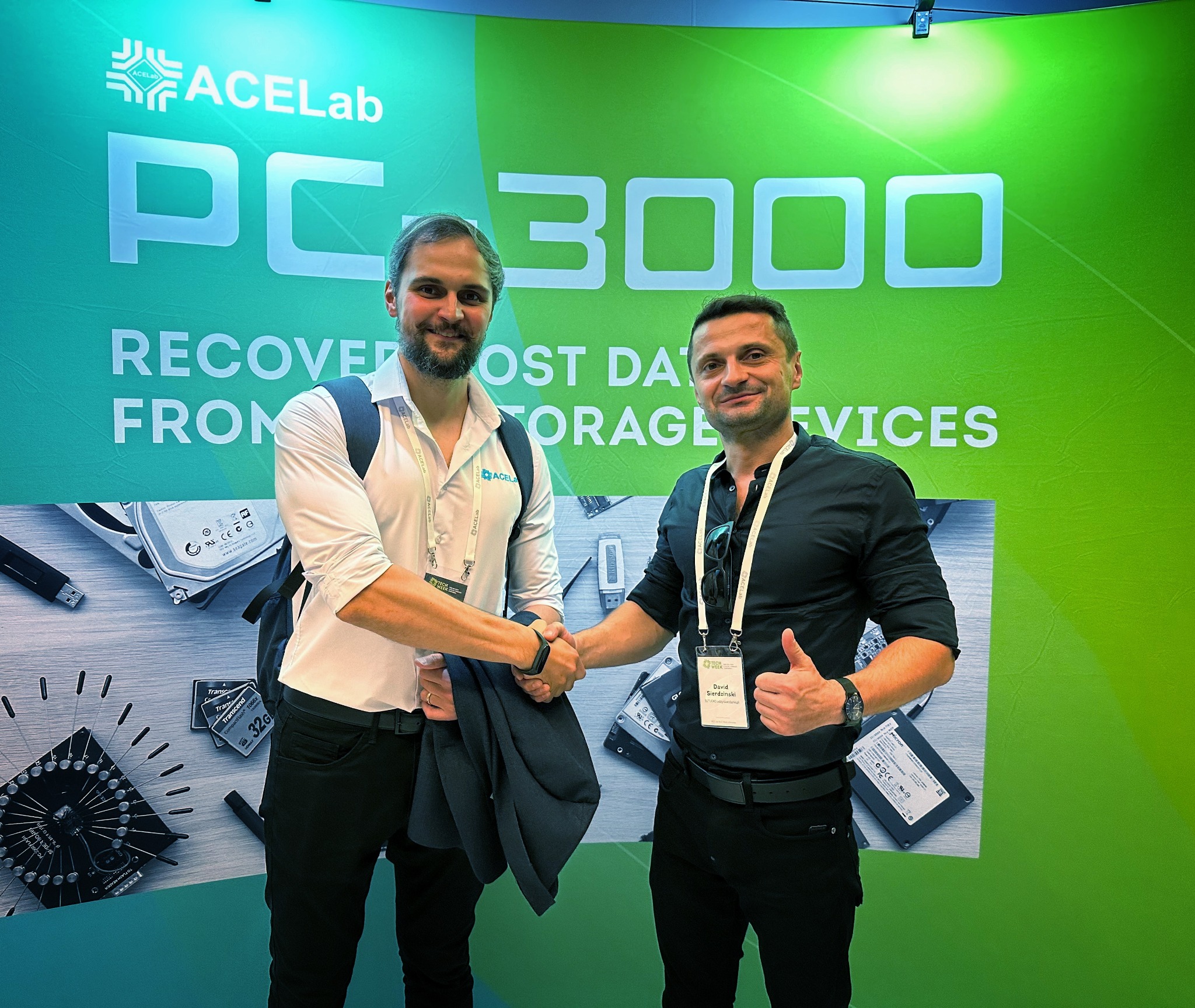 Dwóch mężczyzn wita się na tle stoiska AceLab PC3000 z dyskami i narzędziami do odzyskiwania danych, podczas konferencji branżowej w Maladze. Jeden pokazuje kciuk w górę.
