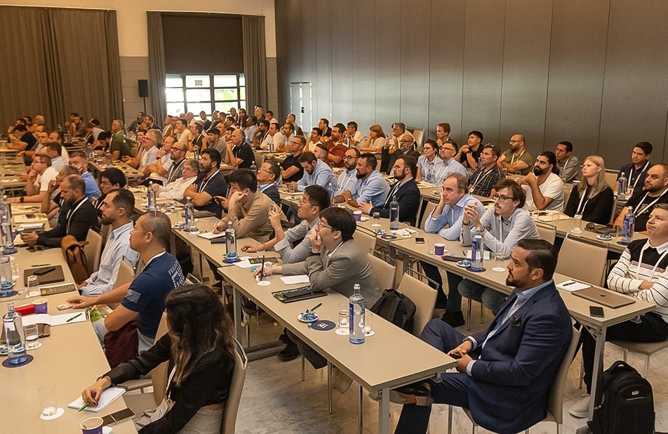 Szeroki kadr sali konferencyjnej z uczestnikami siedzącymi przy stołach, słuchającymi prelekcji. W tle widoczny ekran projekcyjny i okna.