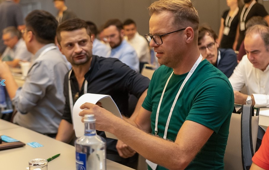 Mężczyzna w okularach analizuje dokument podczas konferencji AceLab. W tle inni uczestnicy, butelka wody i notatki na stole. Koncentracja na wymianie wiedzy.