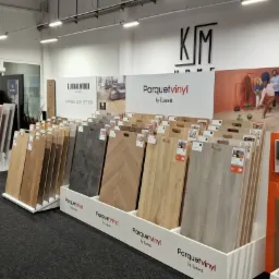 Ekspozycja paneli podłogowych różnych kolorów i wzorów w sklepie, widoczne logo producentów Globalwood i Parquetvinyl by Lamett oraz reklama wodoodporności i łatwości w pielęgnacji.