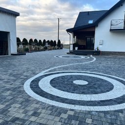 EKO-BUD Rafał Walczak - Efektowna posesja z kostki brukowej w odcieniach szarości, z dekoracyjnymi okręgami. Dom z balkonem i garaż z samochodem w tle, całość w jesiennej scenerii.