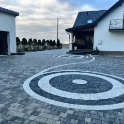 Efektowna posesja z kostki brukowej w odcieniach szarości, z dekoracyjnymi okręgami. Dom z balkonem i garaż z samochodem w tle, całość w jesiennej scenerii.