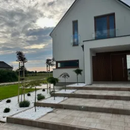 Nowoczesny ogród frontowy z białymi kamieniami, niskimi drzewami i schodami z kostki brukowej prowadzącymi do domu z balkonem. Zachmurzone niebo w tle.