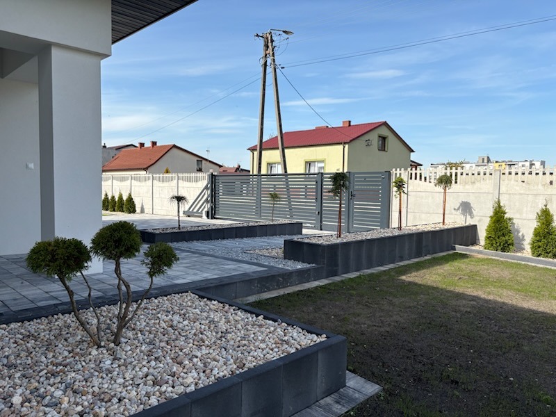 Nowoczesny ogród z szarą kostką brukową, kamiennymi rabatami i ozdobnymi drzewkami, otoczony betonowym ogrodzeniem i metalową bramą. Słoneczny dzień.