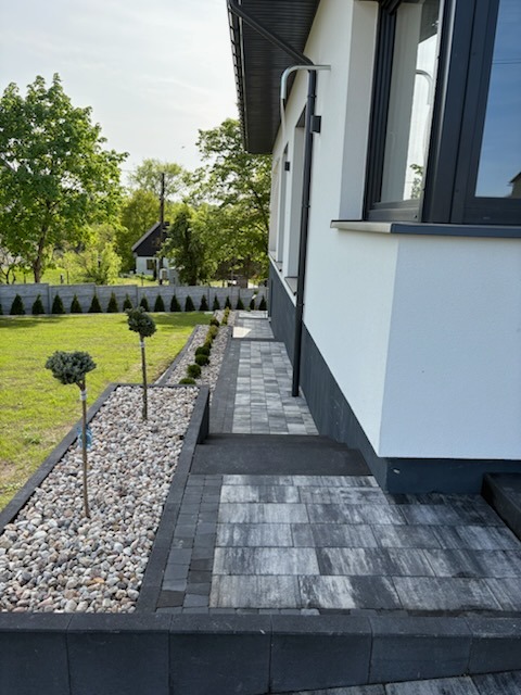 Nowoczesny ogród z ścieżką z kostki brukowej w odcieniach szarości, obrzeża z kamienia i ozdobne drzewka. Estetyczne zagospodarowanie przestrzeni przy domu.