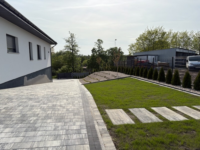 Nowoczesny ogród z kostką brukową w odcieniach szarości, trawnikiem i ścieżką z płyt. W tle budynek z białym tynkiem i zielenią. Widok z perspektywy.