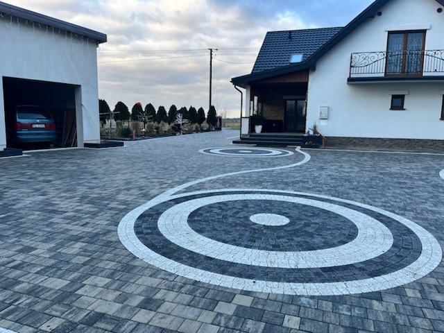 Efektowna posesja z kostki brukowej w odcieniach szarości, z dekoracyjnymi okręgami. Dom z balkonem i garaż z samochodem w tle, całość w jesiennej scenerii.