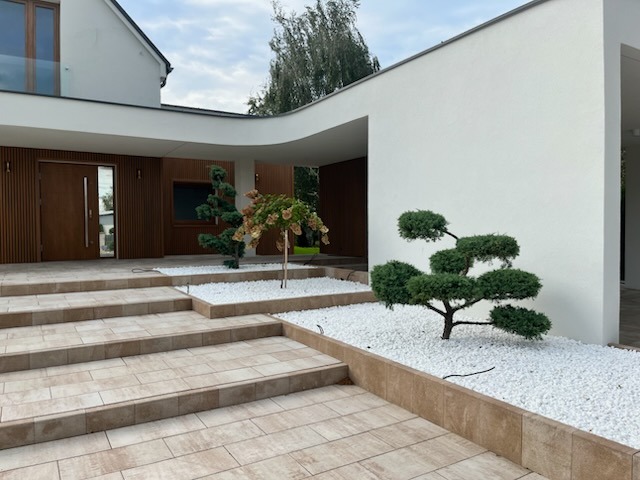 Nowoczesny ogród frontowy z kostki brukowej, białych kamieni i formowanych drzewek. Minimalistyczny design domu z drewnianą fasadą i jasną elewacją. Ścieżka z kostki.