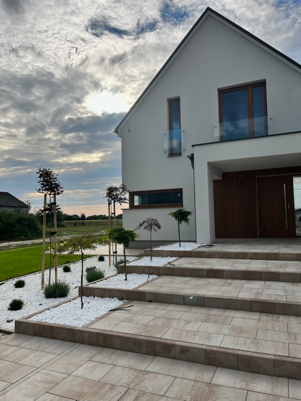 Nowoczesny ogród frontowy z białymi kamieniami, niskimi drzewami i schodami z kostki brukowej prowadzącymi do domu z balkonem. Zachmurzone niebo w tle.