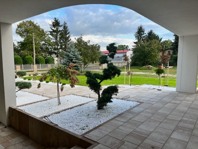 Widok z zadaszonego tarasu na nowocześnie urządzony ogród z białymi kamykami, niskimi drzewkami i równo przystrzyżonym trawnikiem w słoneczny dzień.