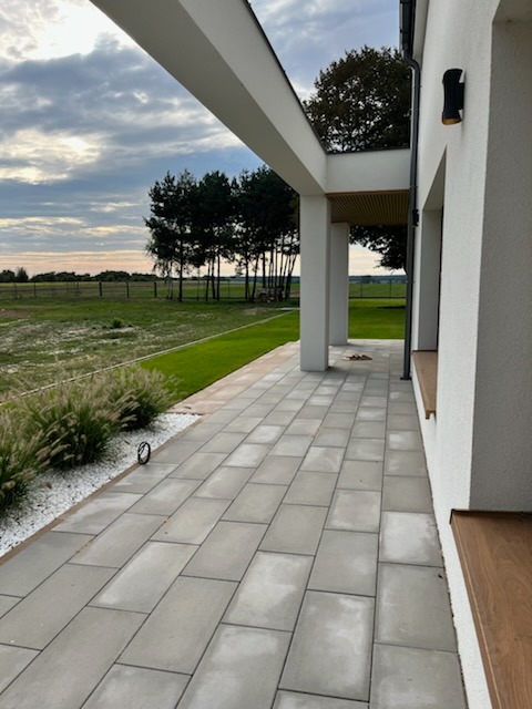 Nowoczesny taras z szarej kostki brukowej, białe kamienie ozdobne, trawnik i widok na łąkę. Minimalistyczny styl, oświetlenie zewnętrzne.