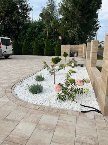 Ogród z kostki brukowej z białymi kamieniami, roślinami i niskim murem. Ułożona kostka brukowa w odcieniach beżu, tworząca podjazd i ścieżkę. Widoczny samochód dostawczy.