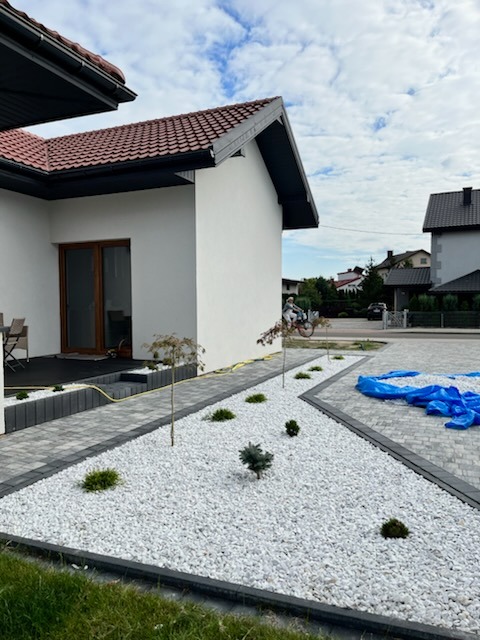 Nowoczesny ogród z białym grysem, ścieżką z kostki i młodymi drzewkami przy jasnym domu. Błękitne niebo w tle, częściowo widoczny chodnik z kostki.