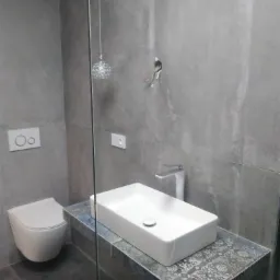 Nowoczesna łazienka z szarą, teksturowaną ścianą, wiszącą toaletą i prostokątną umywalką nablatową na blacie z dekoracyjnym wzorem. Widoczne niedokończone podłączenie elektryczne na ścianie.