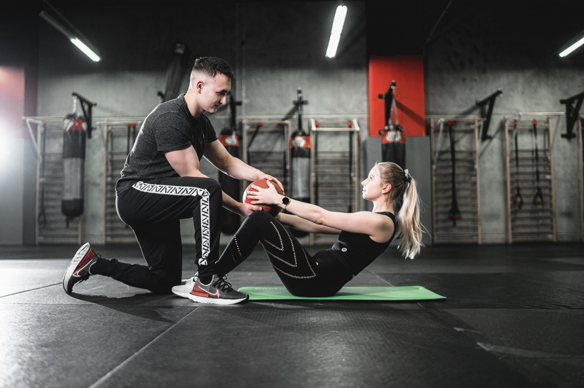 Kobieta wykonuje skręty tułowia z piłką lekarską pod nadzorem trenera personalnego w siłowni z widocznymi workami treningowymi w tle.