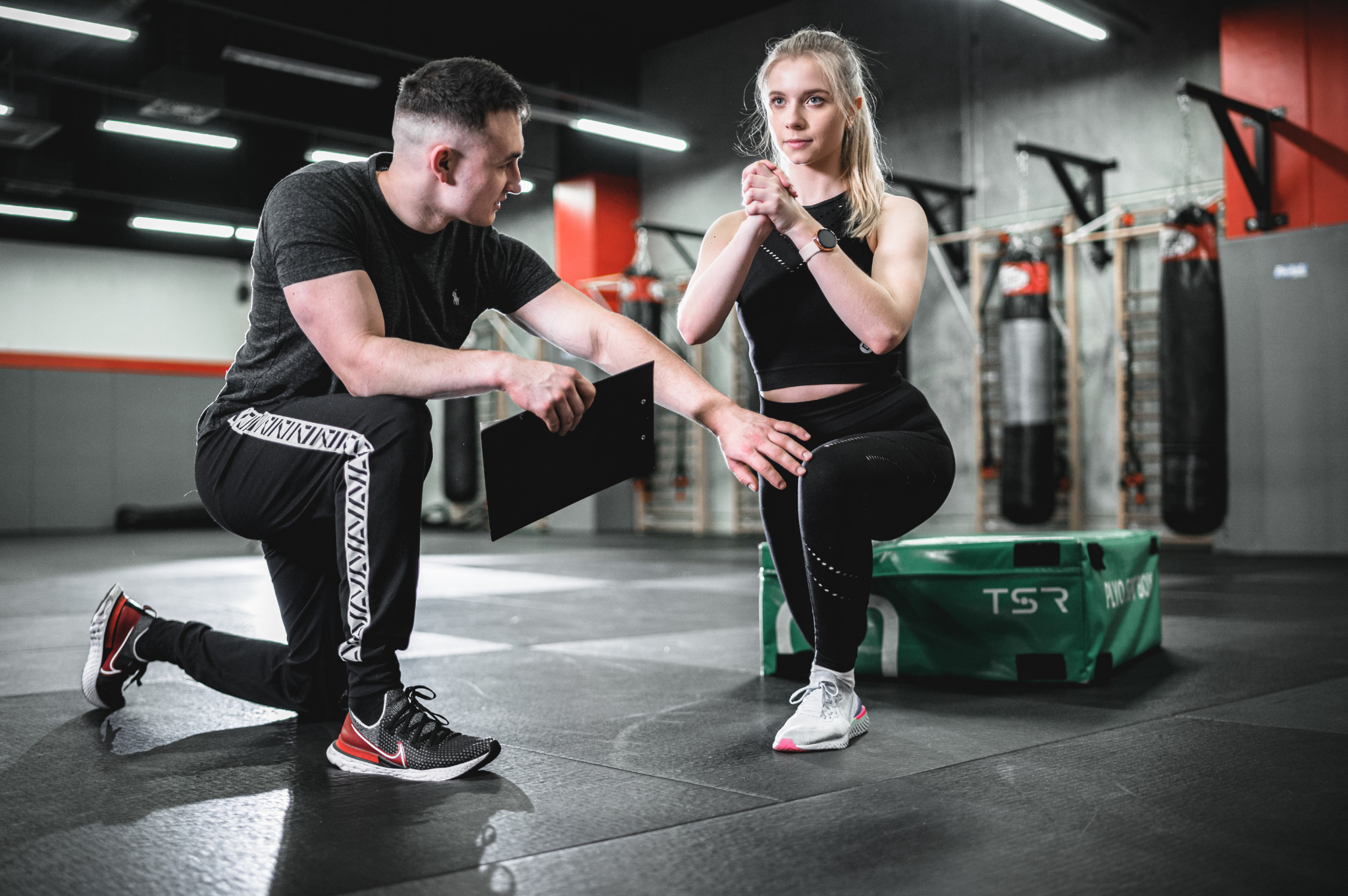 Kobieta w czarnym stroju sportowym wykonuje przysiad z asekuracją trenera w dresowych spodniach i z czarną deską z dokumentami w ręku, na tle worków bokserskich i zielonej skrzyni do ćwiczeń.