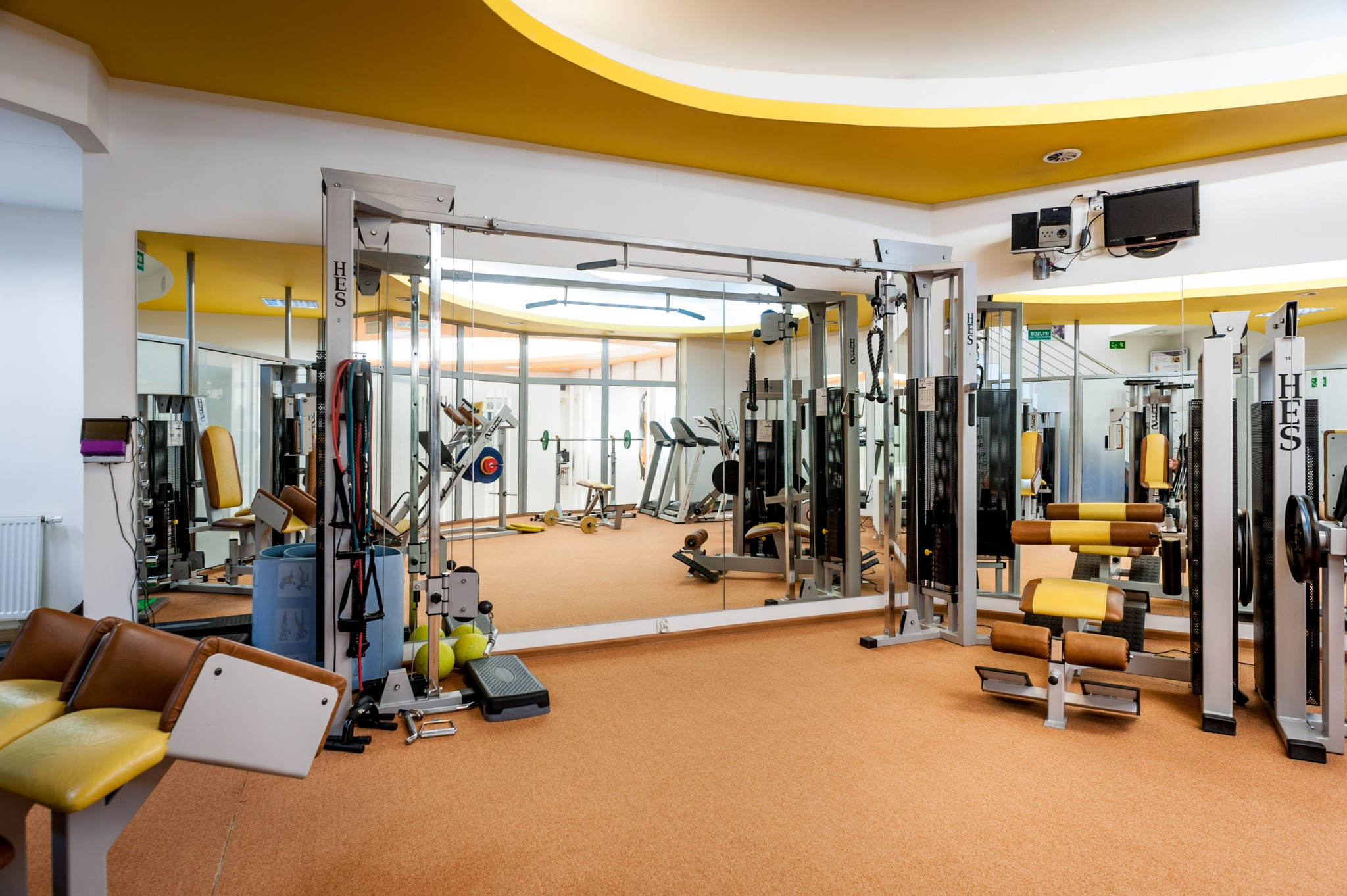 Jasne wnętrze siłowni z lustrami, widoczne maszyny do ćwiczeń, bieżnie, ciężary i akcesoria do treningu, żółte akcenty w oświetleniu sufitowym.