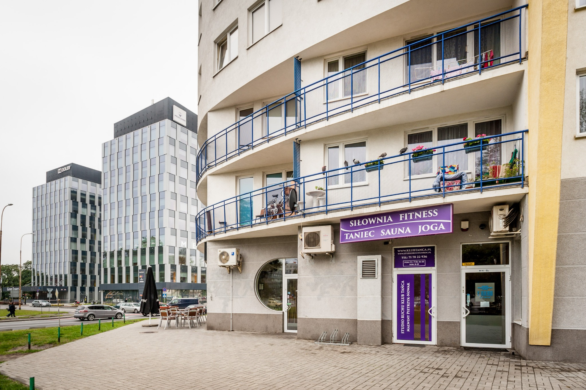 Wejście do studia ruchu z fioletowymi akcentami, szyld reklamujący siłownię, fitness, taniec, saunę i jogę, budynek z zaokrąglonymi balkonami, w tle nowoczesne biurowce.