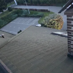 Dach pokryty bitumiczną dachówką z posypką, widoczny komin z cegły, okna dachowe i fragment rynny, w tle parking i zieleń.