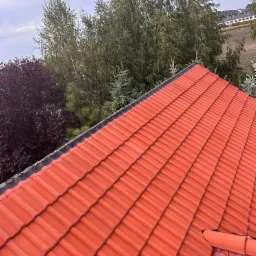 Czerwona dachówka ceramiczna na skośnym dachu domu jednorodzinnego, widok z góry, w tle drzewa i zabudowa mieszkalna.