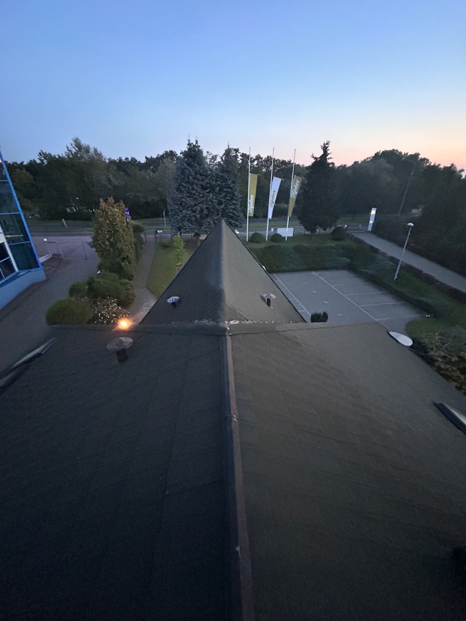 Widok z góry na ciemny, płaski dach z kominem, z widoczną okoliczną zielenią i elementami architektury w tle, sfotografowany o zmierzchu.