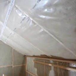 Izolacja poddasza wełną mineralną, zabezpieczona folią paroizolacyjną z nadrukiem producenta, widoczna drewniana konstrukcja szkieletowa dachu.