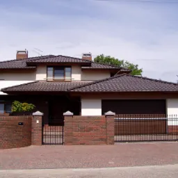 Dwukondygnacyjny dom z brązowym dachem, jasną elewacją i ceglanym ogrodzeniem z metalową bramą, widziany z poziomu ulicy w słoneczny dzień.