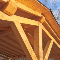 Drewniana konstrukcja z belek i desek, widok od dołu, jasne drewno, widoczne połączenia elementów, w tle drzewa i niebieskie niebo.