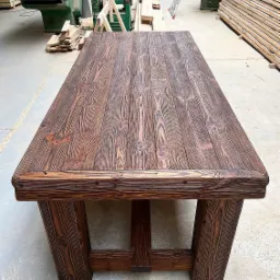 Masywny, rustykalny stół z ciemnego drewna sosnowego, w tle warsztat stolarski z maszynami i ułożonymi deskami.