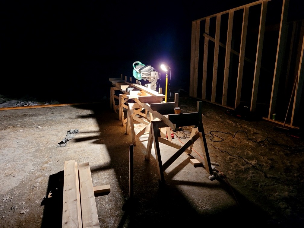 Konstrukcja szkieletowa domu w trakcie budowy nocą, oświetlona lampą budowlaną, z widoczną piłą stołową na drewnianym stole roboczym i stertą desek na ziemi.