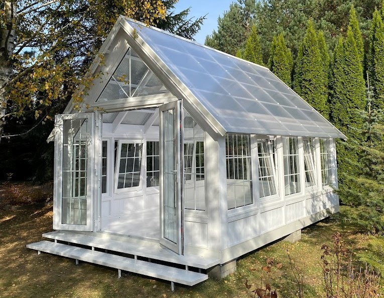 Biały, przeszklony budynek z przezroczystym dachem, otoczony zielenią i drzewami, z otwartymi dwuskrzydłowymi drzwiami i schodkami wejściowymi.