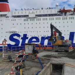 Żółta koparka Volvo na nabrzeżu portowym, w tle duży biały prom pasażerski Stena Line, robotnicy w pomarańczowych kamizelkach, wykopane kable.