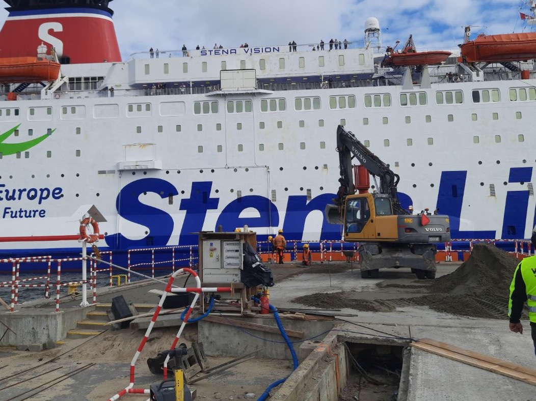 Żółta koparka Volvo na nabrzeżu portowym, w tle duży biały prom pasażerski Stena Line, robotnicy w pomarańczowych kamizelkach, wykopane kable.
