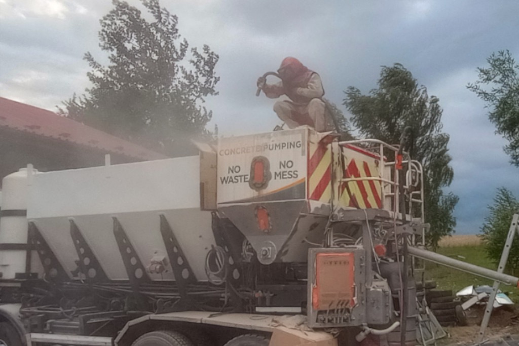 Pracownik w kombinezonie ochronnym piaskuje silos na betoniarce, z widocznym napisem 'Concrete Pumping', 'No Waste', 'No Mess', na zewnątrz, z drzewami w tle.