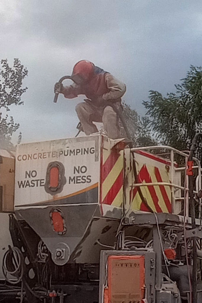 Pracownik w kombinezonie ochronnym i masce, siedzący na zbiorniku betonomieszarki, wykonuje prace czyszczące strumieniem ścierniwa.