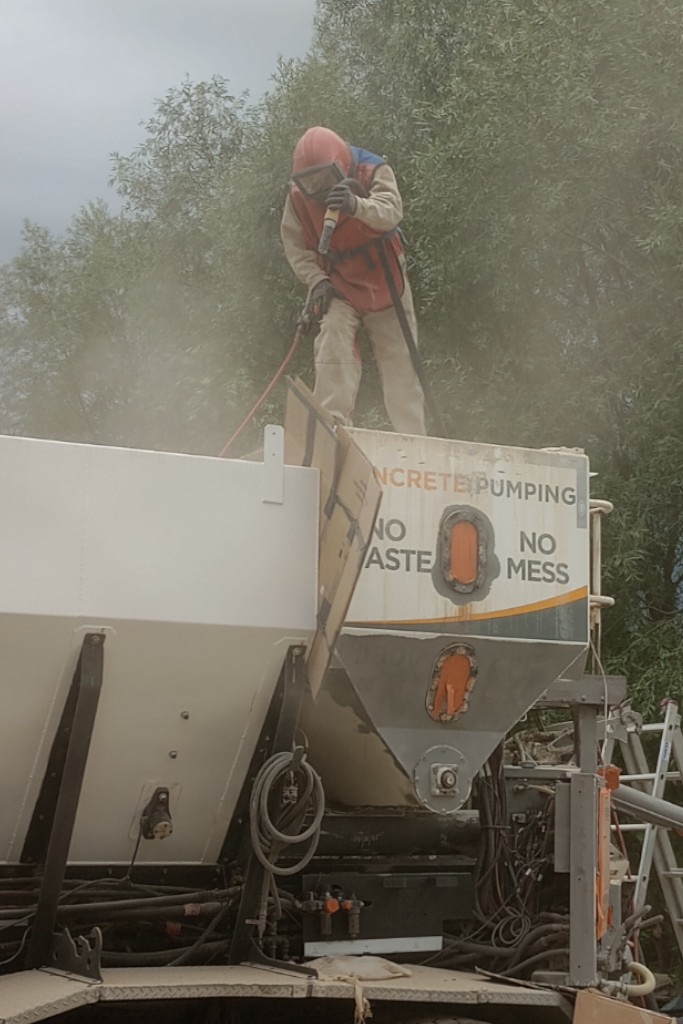 Pracownik w kombinezonie ochronnym i masce piaskuje białą naczepę betoniarki z napisem 'CONCRETE PUMPING NO WASTE NO MESS', unoszący się pył w powietrzu, drabina w tle.