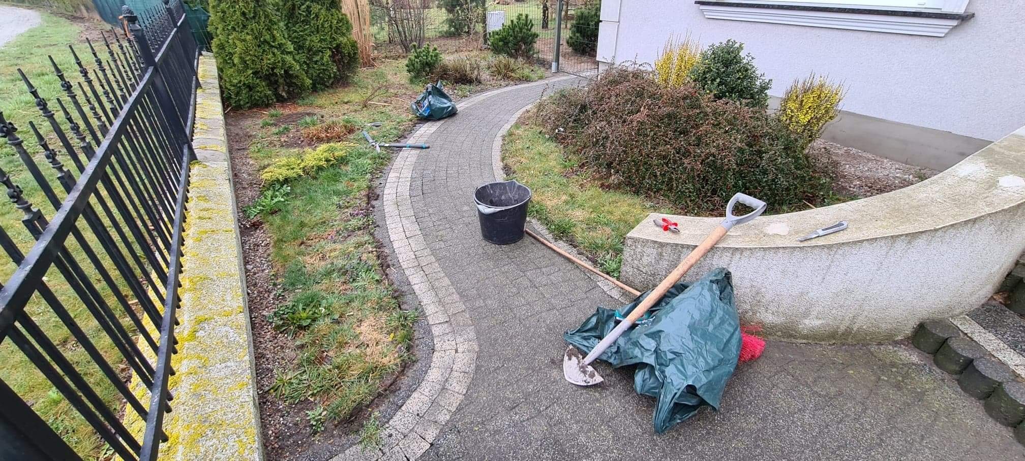 Ścieżka z kostki brukowej w ogrodzie w trakcie porządkowania, widoczne narzędzia ogrodnicze, wiadro i worki na odpady roślinne.