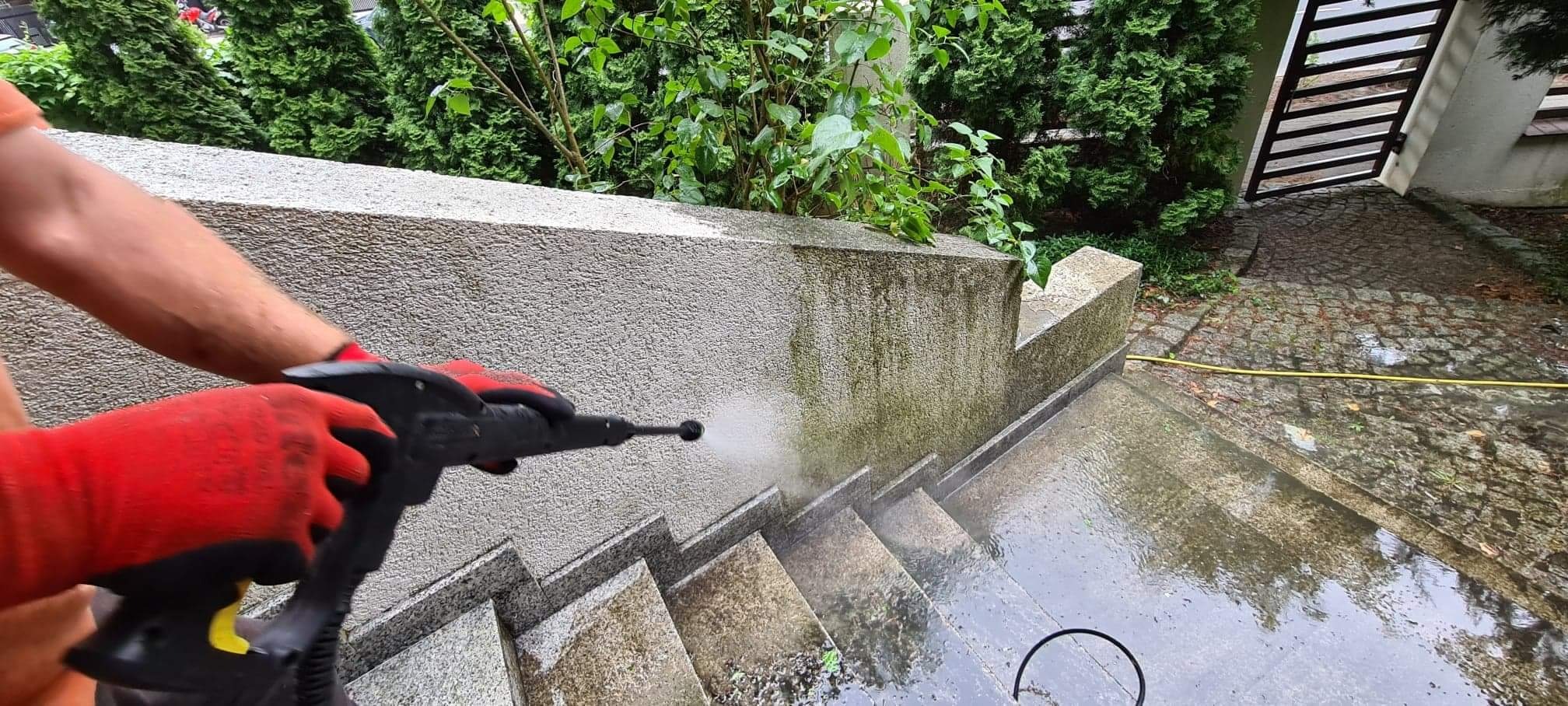 Czyszczenie kamiennych schodów myjką ciśnieniową, widoczne czerwone rękawice ochronne i strumień wody usuwający zabrudzenia, w tle zielona roślinność i brukowana ścieżka.