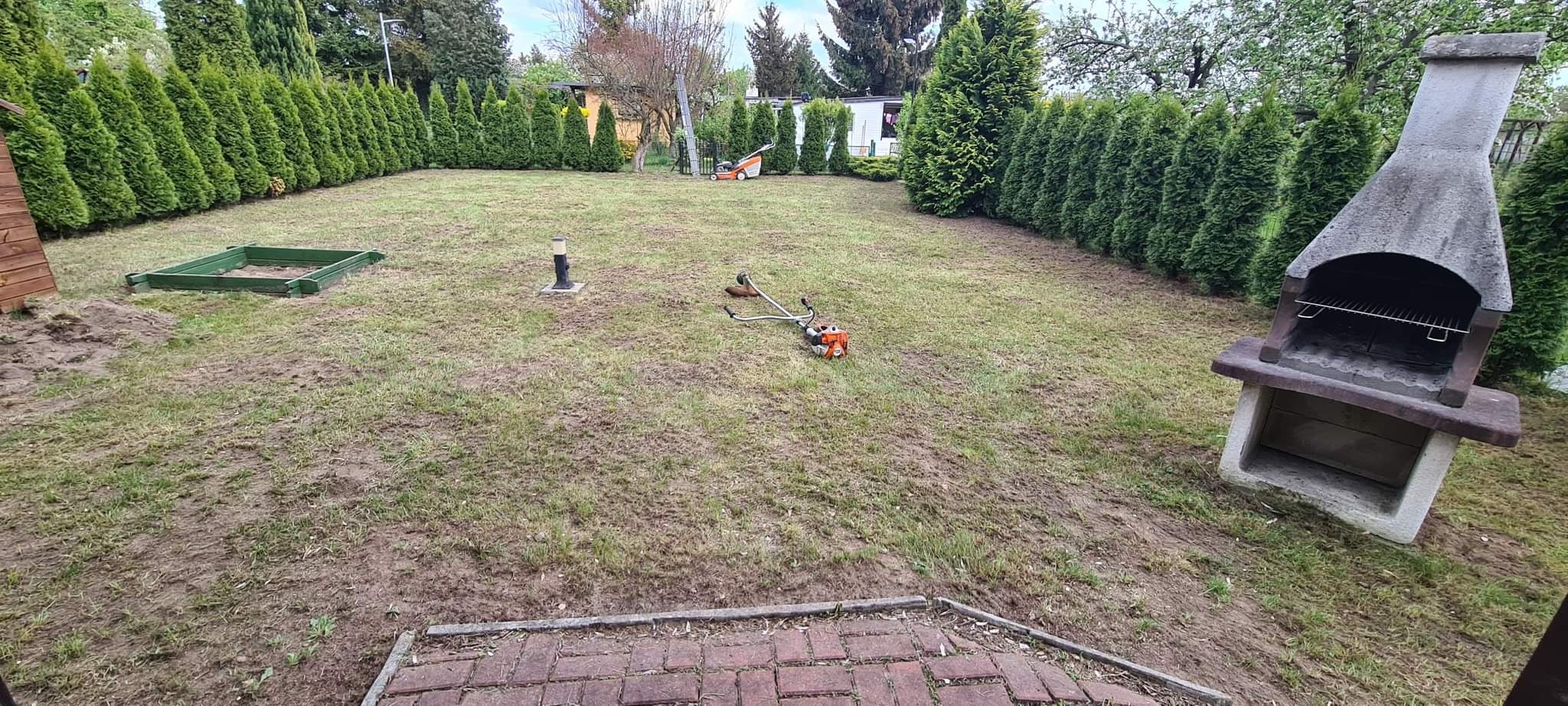 Przygotowanie terenu pod trawnik: glebogryzarka i podkaszarka leżą na ziemi, w tle rząd tuj, piaskownica i murowany grill.