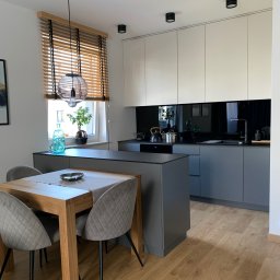 AKdecor - Studio projektowe - Nowoczesna kuchnia z wyspą, drewnianym stołem i szarymi krzesłami, oświetlona lampą wiszącą i światłem wpadającym przez okno z drewnianymi żaluzjami.