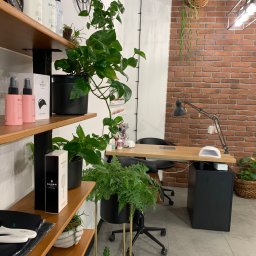 AKdecor - Studio projektowe - Wnętrze gabinetu kosmetycznego z drewnianym biurkiem, lampą, roślinami doniczkowymi, półkami z kosmetykami Balmain i ścianą z cegły.