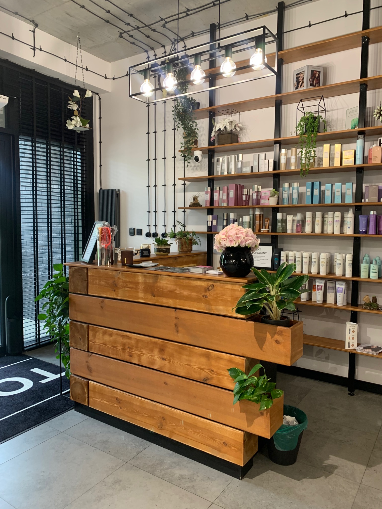 Nowoczesne wnętrze salonu fryzjerskiego z drewnianą recepcją, półkami z kosmetykami i dekoracyjnymi roślinami doniczkowymi.