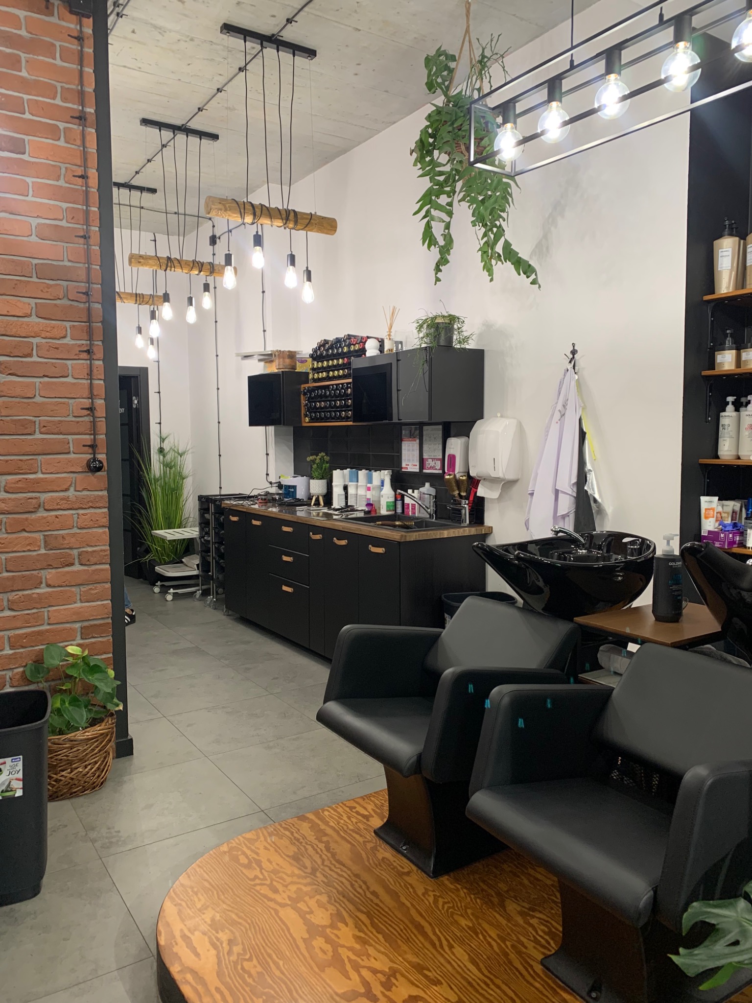 Nowoczesne wnętrze salonu fryzjerskiego z czarnymi meblami, drewnianymi akcentami, ceglaną ścianą i roślinami wiszącymi pod sufitem, ukazujące stanowiska do mycia włosów i szafki z produktami.