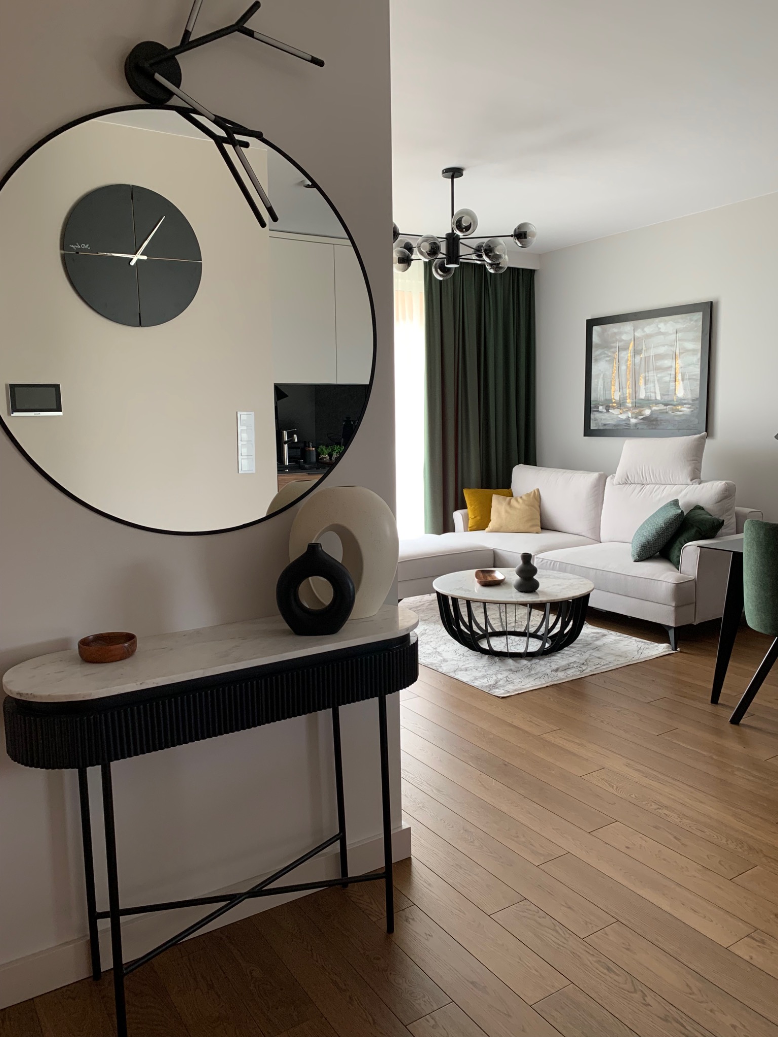 Nowoczesny salon z szarą sofą narożną, okrągłym stolikiem kawowym z marmurowym blatem i dekoracyjnym lustrem z zegarem nad konsolą z wazonami.