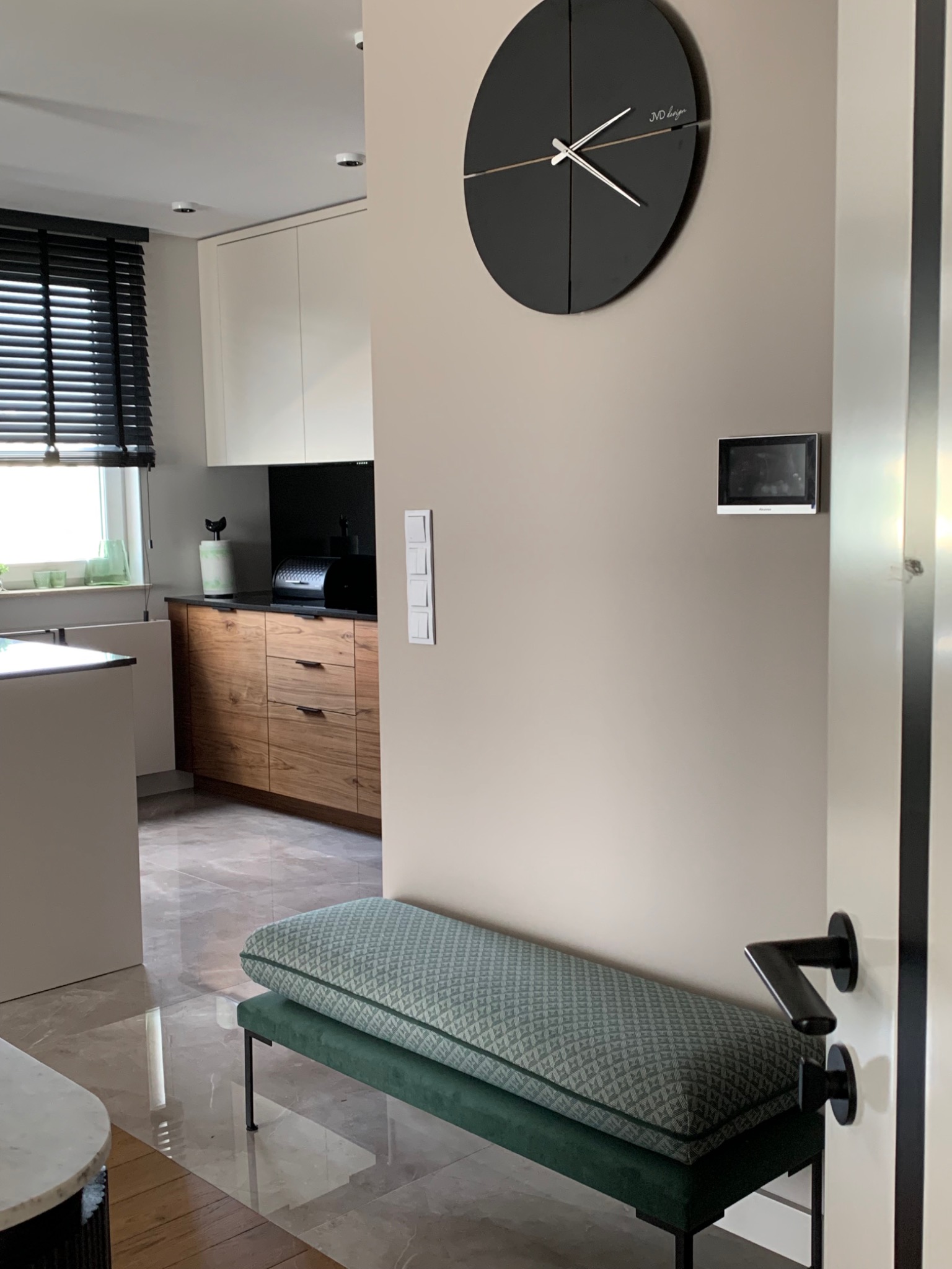 Nowoczesne wnętrze: ławka z zieloną tapicerką w przedpokoju, widok na kuchnię z drewnianymi frontami i czarnymi żaluzjami, duży czarny zegar na ścianie.