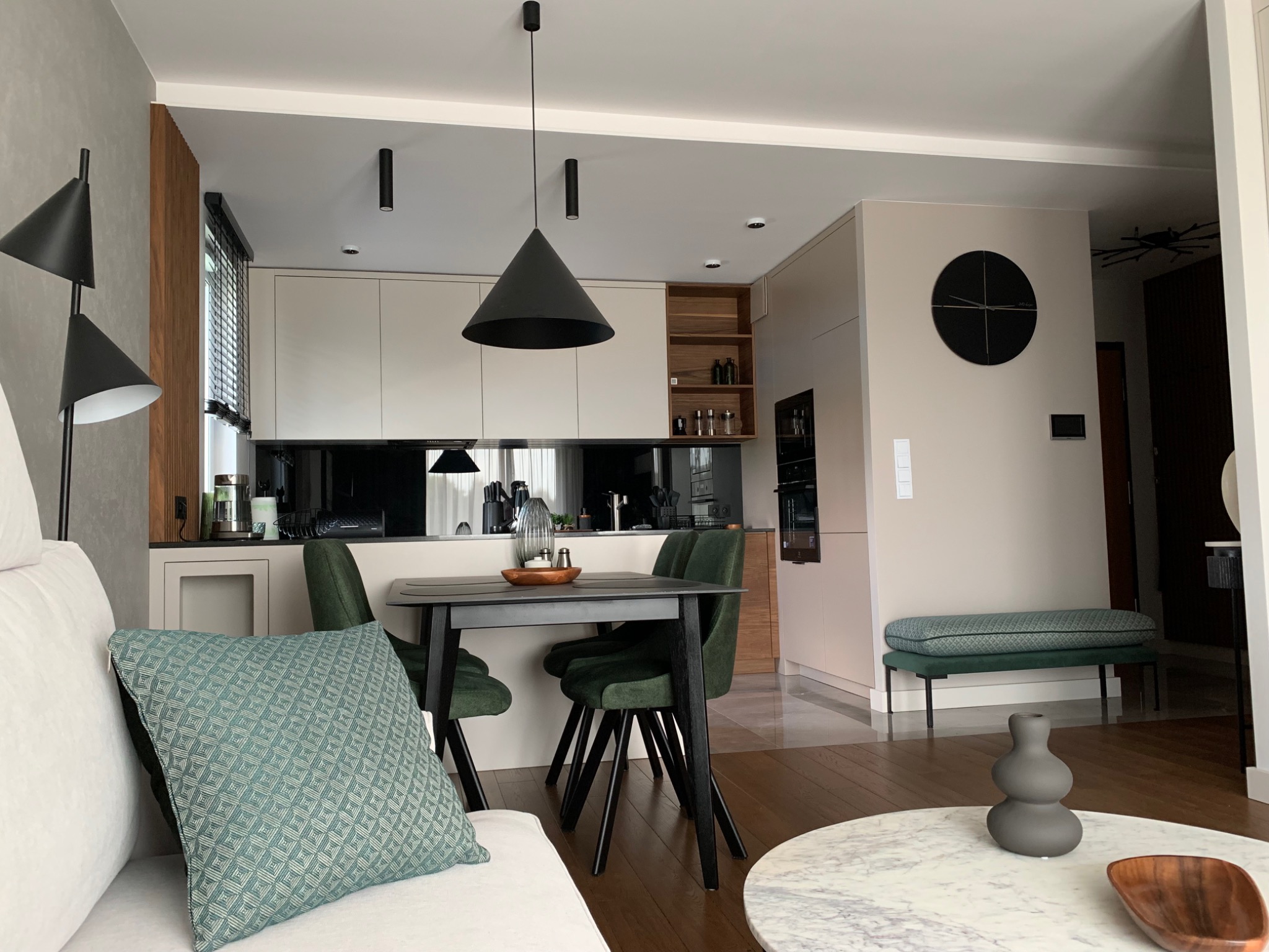 Nowoczesne wnętrze mieszkania z otwartą kuchnią, widoczny stół z zielonymi krzesłami, sofa z poduszką w geometryczny wzór oraz okrągły stolik kawowy z marmurowym blatem.