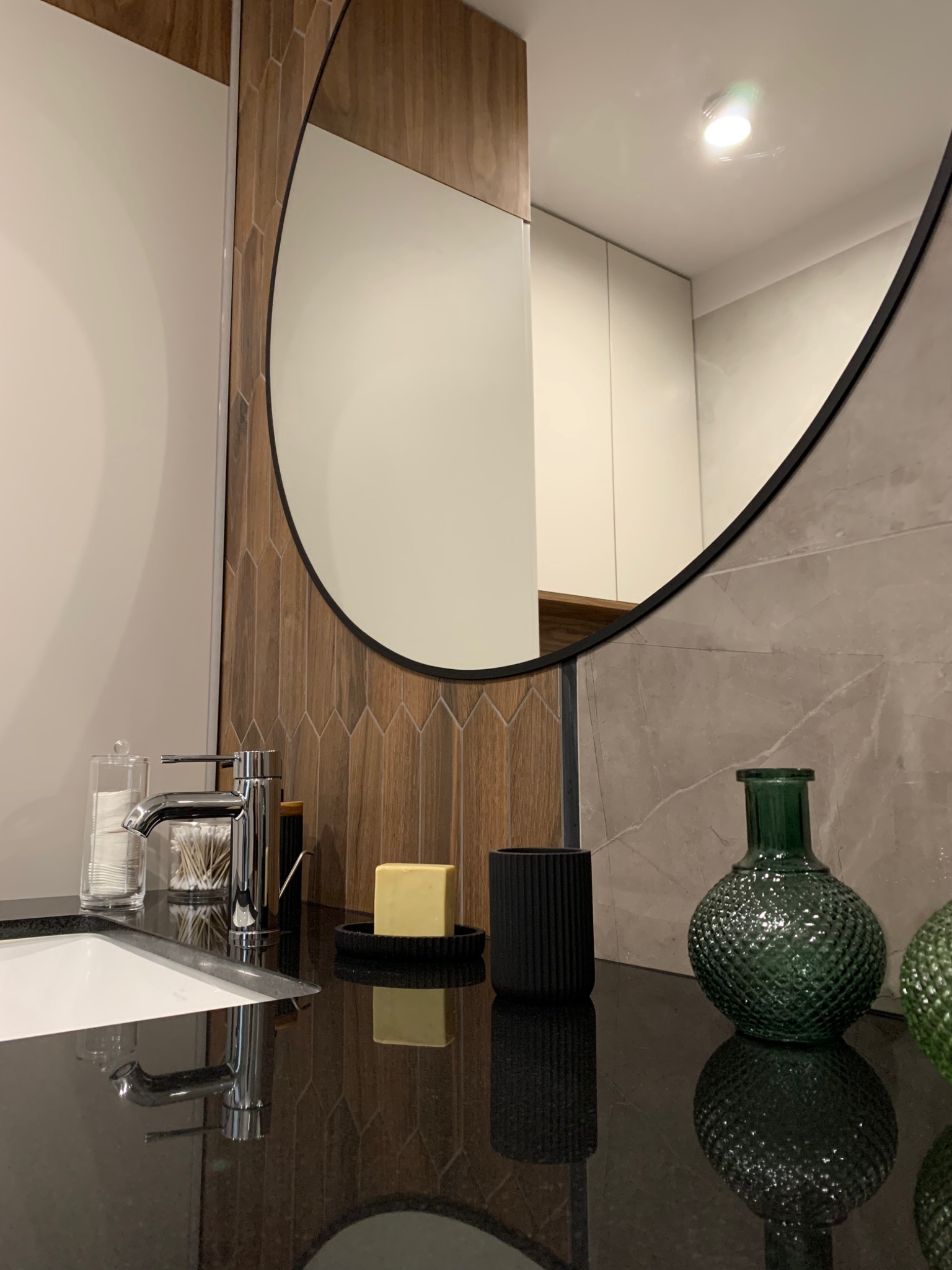 Nowoczesna łazienka z okrągłym lustrem, chromowaną baterią, drewnopodobnymi płytkami ułożonymi w jodełkę i zielonymi szklanymi wazonami na czarnym blacie.