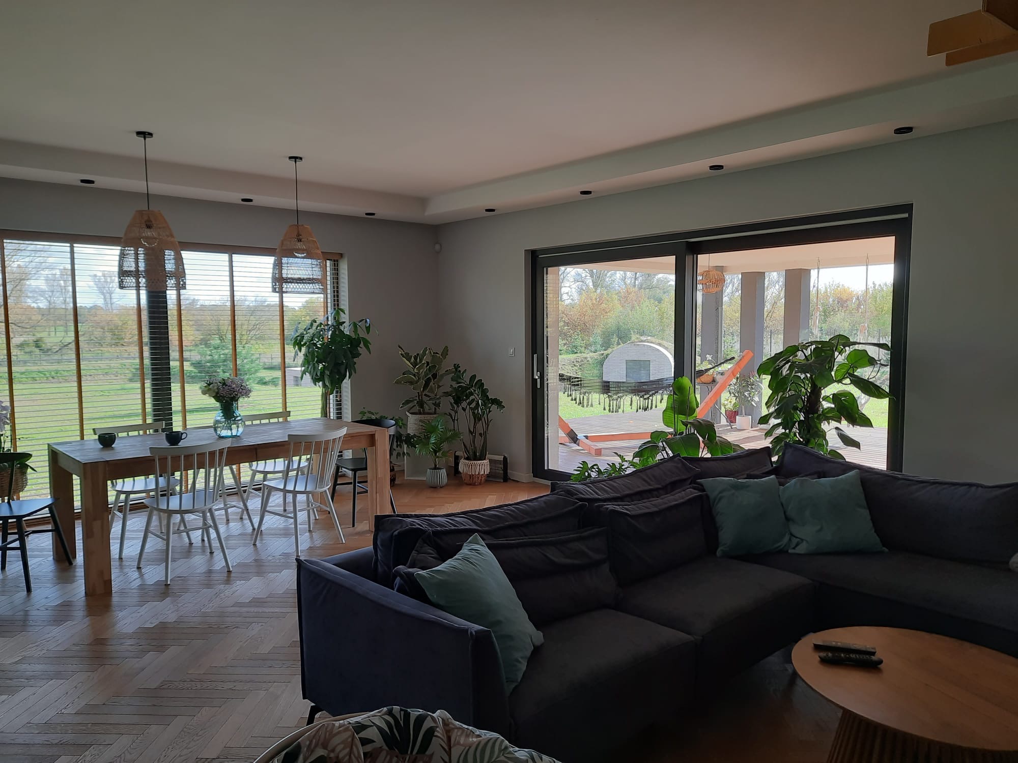 Jasny salon z drewnianą podłogą w jodełkę, dużym narożnikiem z poduszkami, drewnianym stołem z białymi krzesłami przy oknie z żaluzjami i roślinami doniczkowymi.