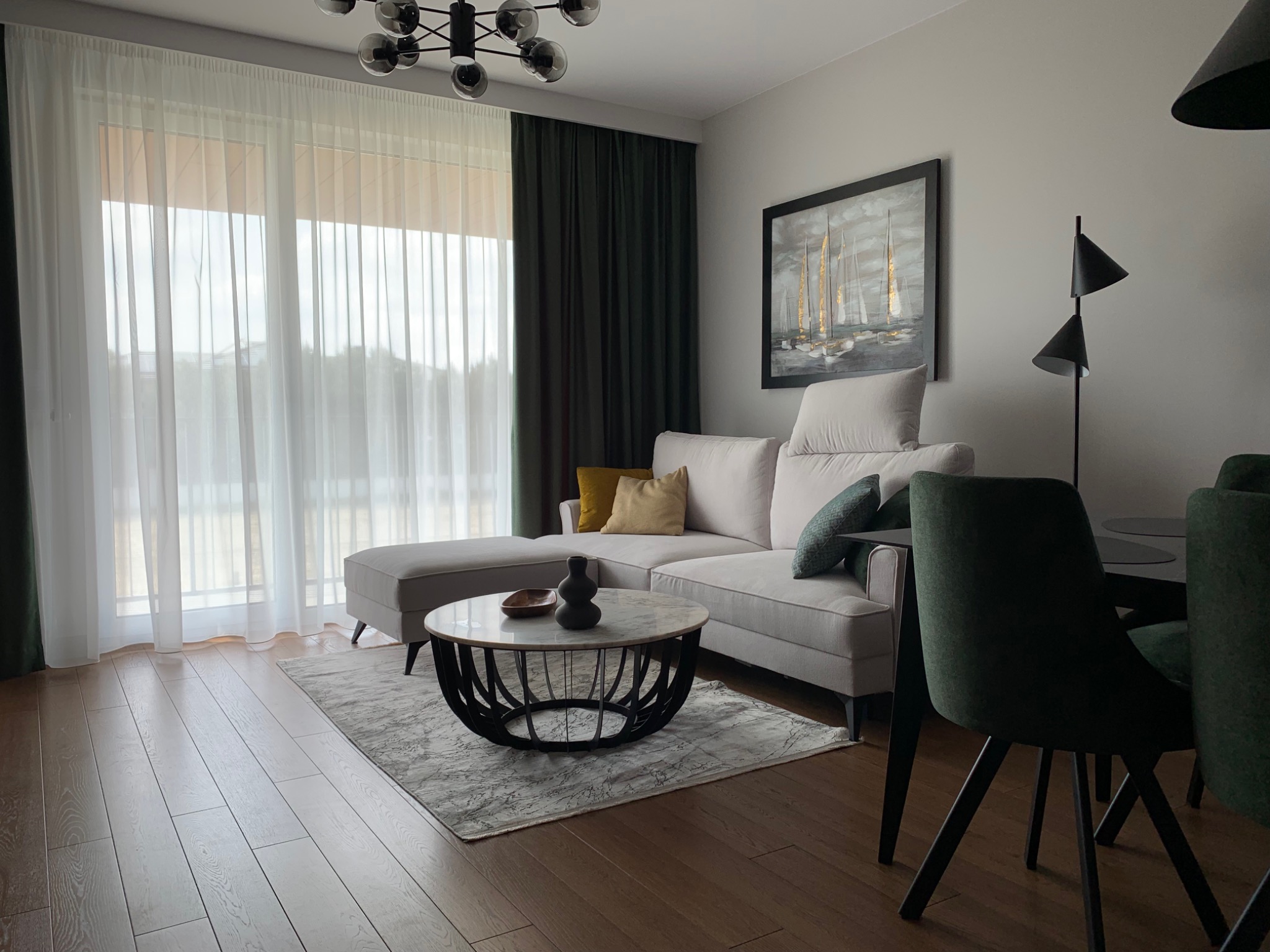 Jasny salon z narożną sofą, okrągłym stolikiem kawowym z marmurowym blatem i ciemnozielonymi akcentami w postaci zasłon i krzeseł, obraz z motywem żaglówek na ścianie.