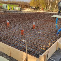 Zbrojenie płyty fundamentowej w drewnianym szalunku, widoczne pionowe pręty, rury kanalizacyjne, czarna folia izolacyjna i dwóch pracowników na budowie.